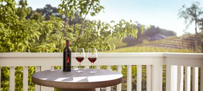 White French doors open to a patio with two wooden chairs, two glasses of red wine, and a view of the vineyard hills. 