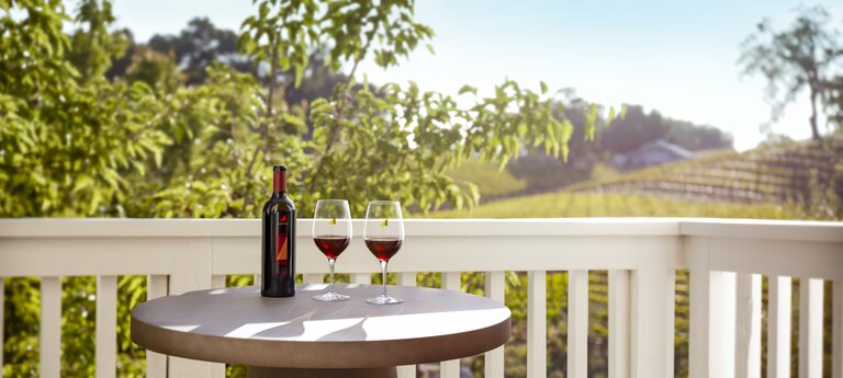 White French doors open to a patio with two wooden chairs, two glasses of red wine, and a view of the vineyard hills.  