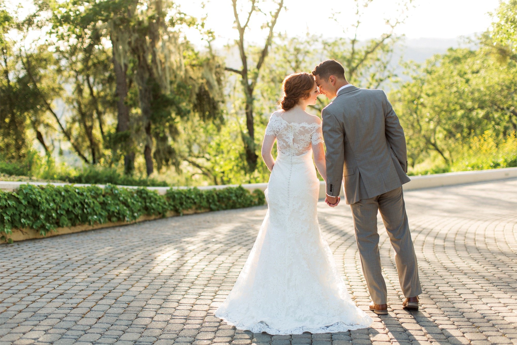 A bride and groom walk down a cobblestone road toward vibrant greenery, holding hands and leaning in to kiss. 