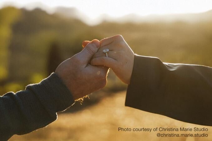 A bride and groom walk down a cobblestone road toward vibrant greenery, holding hands and leaning in to kiss. 