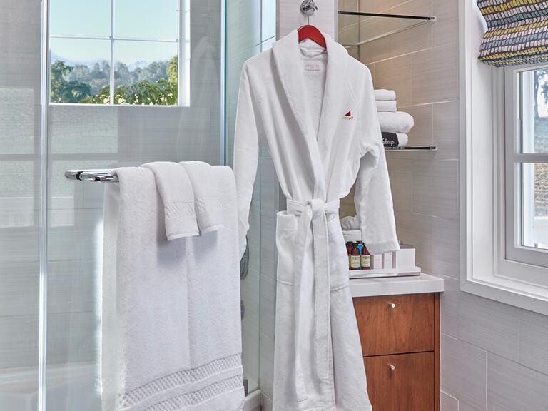 A bathroom with beige tile walls, a full shower with a window above it on the left, complete with towels and a bathrobe, followed by a shelf and a window to the right.