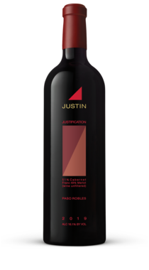 A bottle of JUSTIFICATION standing in front of a pink and purple sunset over the vineyard hills. 