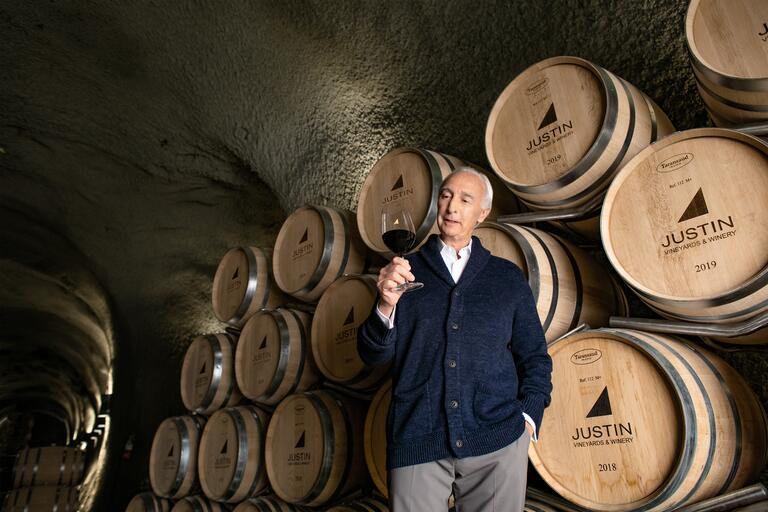 Justin Baldwin describes the glass of red wine in his hand, standing casually in front of JUSTIN 2019 wooden barrels.