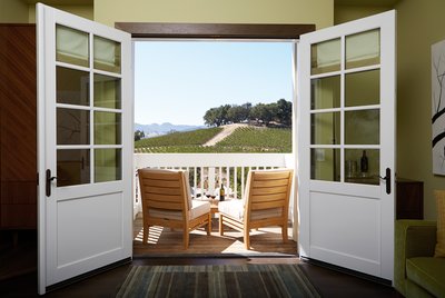 White French doors open to a patio with two wooden chairs, two glasses of red wine, and a view of the vineyard hills. 