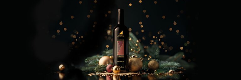 A bottle of JUSTIN Isosceles surrounded by red and gold Christmas ornaments, pine greenery, and gold sparkles on a black background.