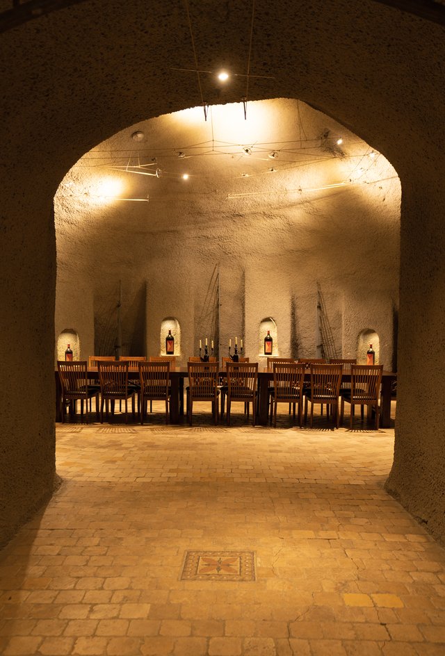 A long table sits in a domed, cement room, alcoves in the walls displaying bottles of ISOSCELES with a soft glow.