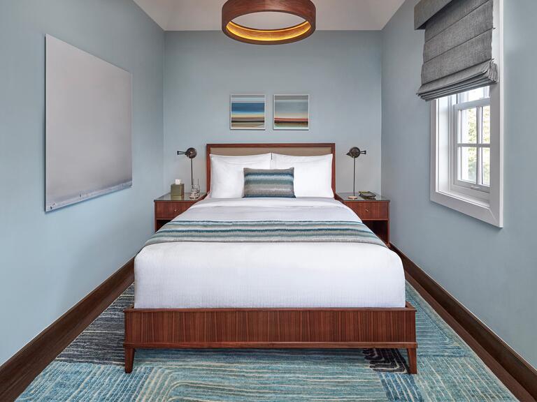 A bedroom with slate blue walls, complete with a queen-sized bed with two small pictures above it, matching wooden bedside tables, and a window on the right