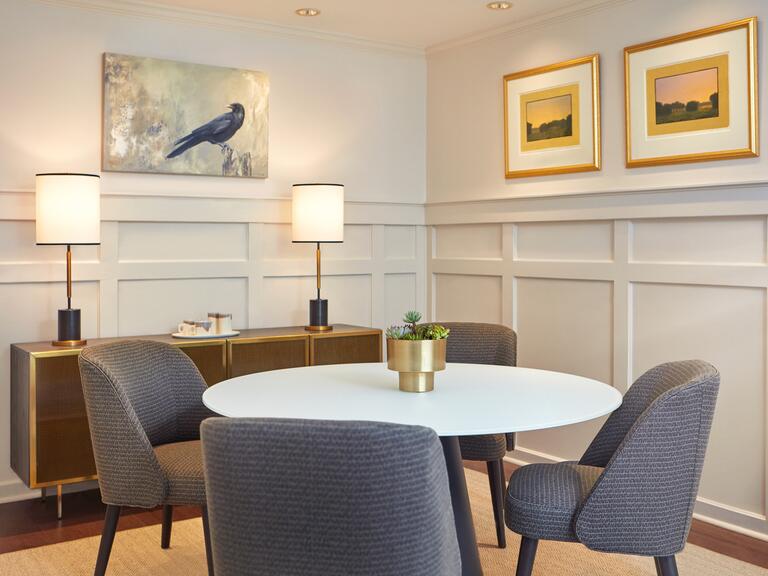 A dining area with white walls, a large brown and gold console table to the left, a white circular table with four blue chairs, and artwork on both walls