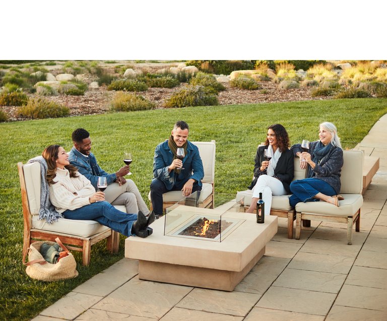 In the late afternoon light, five friends lounge around a firepit, smiling and enjoying glasses of ISOSCELES RESERVE.