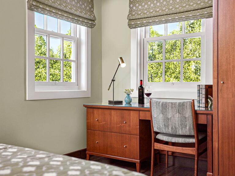 Wooden desk facing two large windows overlooking trees. 