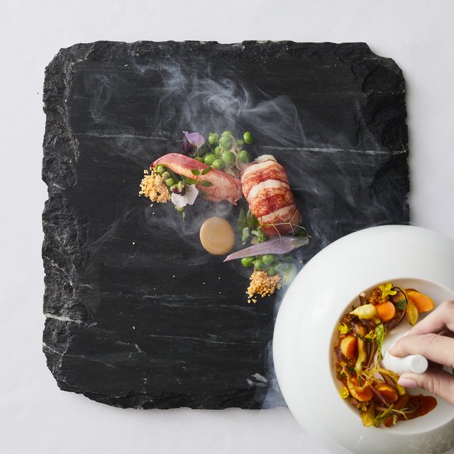 A black slab of stone with shrimp, peas, and flowers, enveloped in steam coming from a bowl of colorful morsels. 