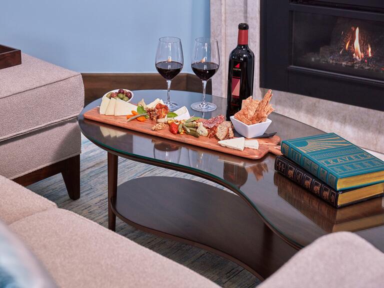A living room with light blue walls and a fireplace, containing midcentury modern wooden furniture with the photo's primary focus being a coffee table where a charcuterie board, wine, and books lay