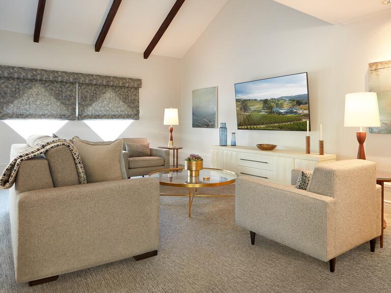 A living room with white walls with grey furniture, diamond-shaped windows, a golden coffee table, and a large white tv console along with a tv and two paintings on both sides
