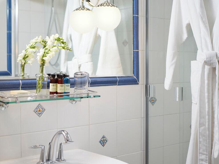 A closeup of a mirror and glass shelf above a ceramic sink and a shower to the left, complete with a bathrobe
