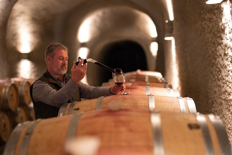 JUSTIN Winemaker Greg Stach injects red wine into a glass with a barrel sampler in the barrel-stacked, cement caves.