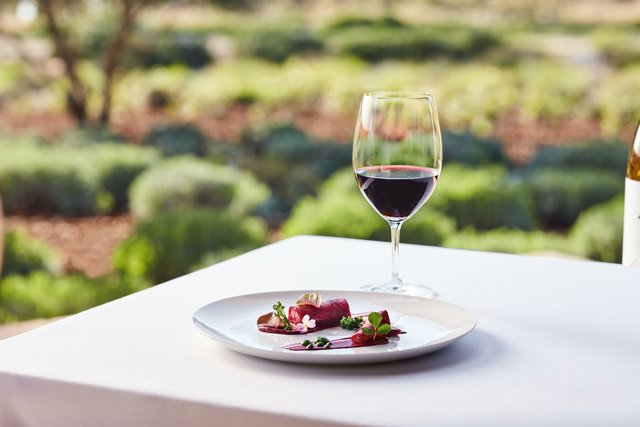 A glass of red wine and a plate of vivid beet salad garnished with greens sit on a white table overlooking the vineyard.