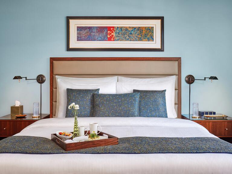 A bedroom with teal walls adorned with a long, horizontal picture above a queen-sized bed complete with two matching wooden, bedside tables on each side
