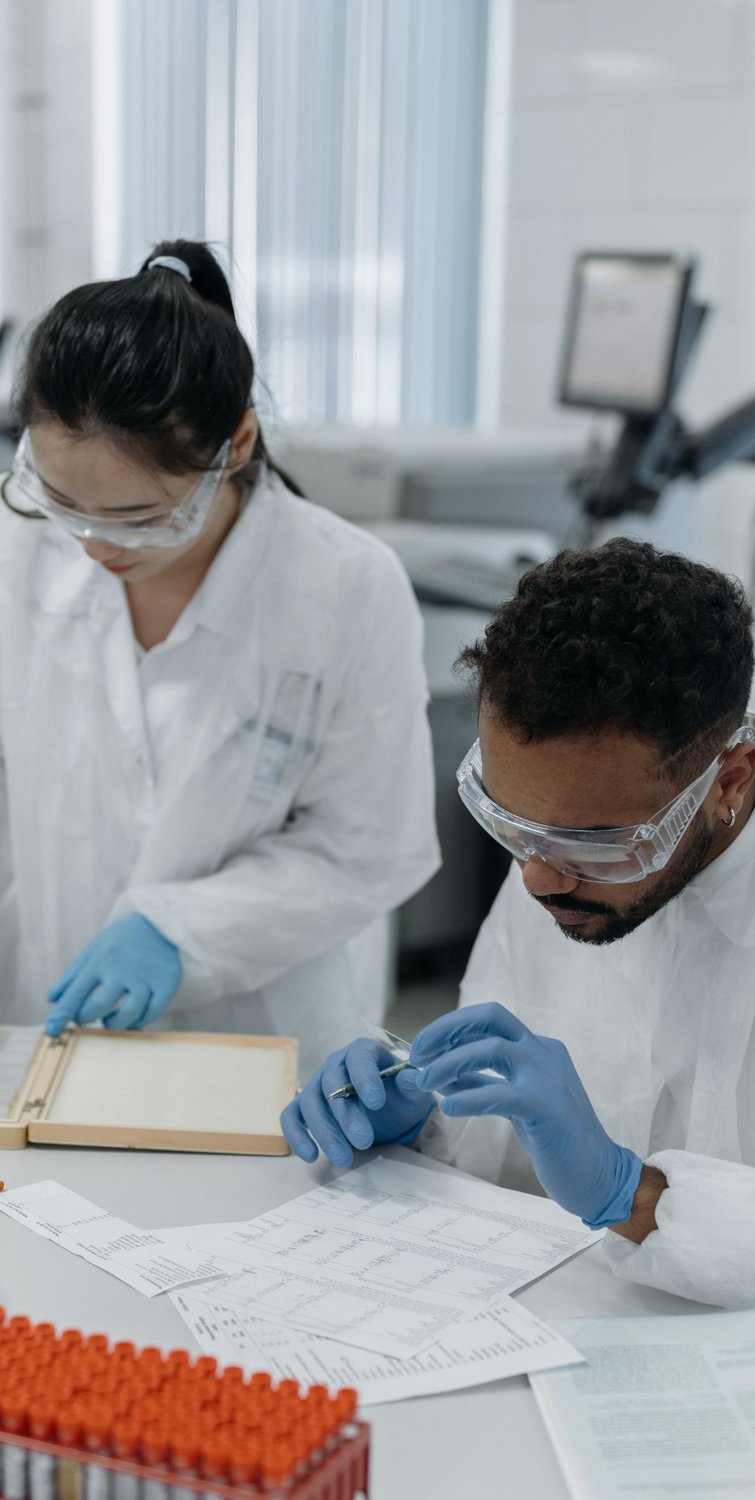 Two scientists in white lab coats with blue gloves examining some papers on a table in a lab