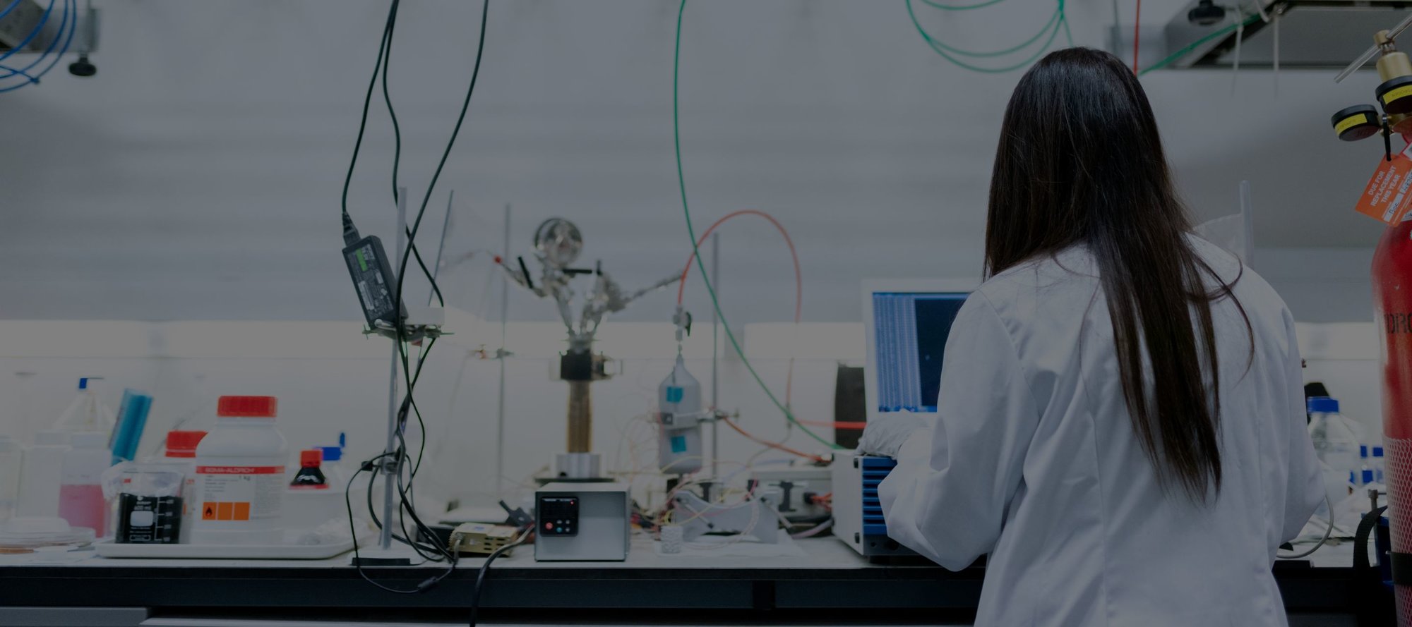 A scientist in a lab coat viewed from behind working on a computer surrounded by tubes and equipment