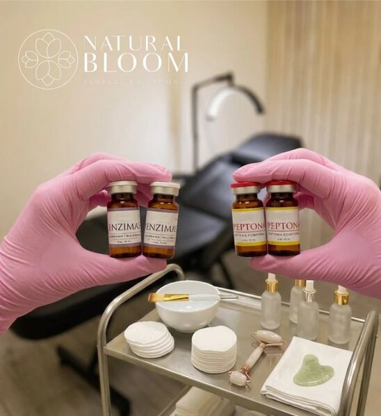 Manos con guantes rosa sosteniendo viales de enzimas y peptonas sobre una mesa auxiliar con herramientas de skincare facial en una clínica estética Natural Bloom.