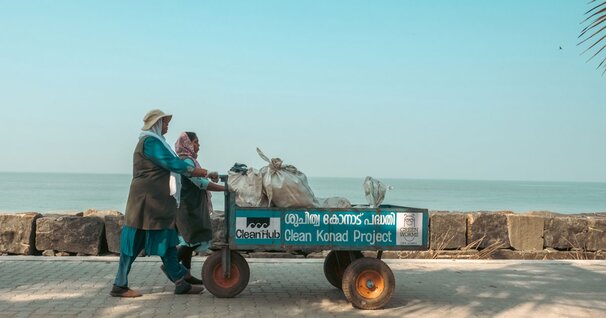 Images shows a pristine coastal area, with two superstars pushing a waste collection cart, carrying bags of waste collected from houses, instead of being dumped near waterways.