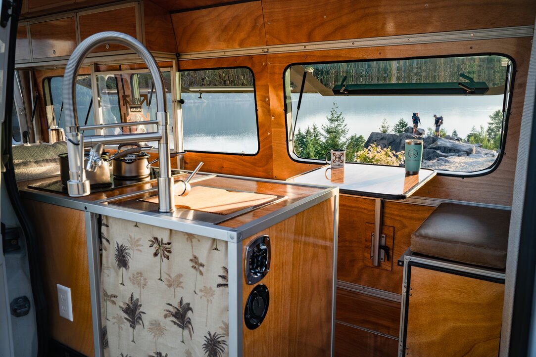 A woman enjoying a morning in her camper van kitchen, pouring coffee while admiring the ocean view. Experience serenity and convenience in our camper van.