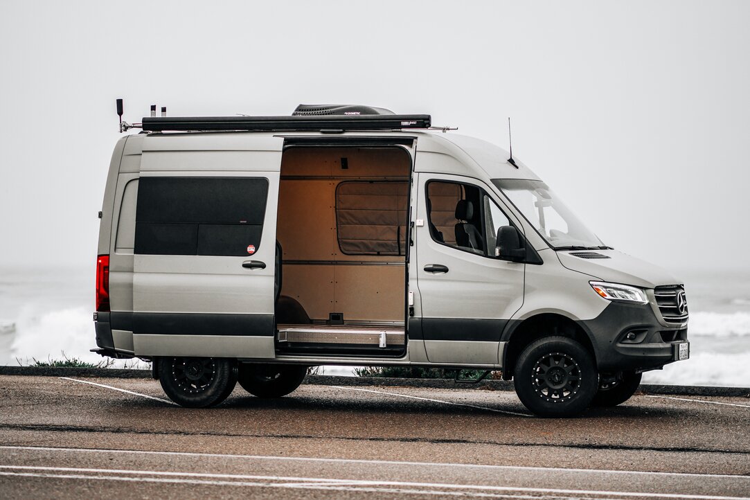 A woman enjoying a morning in her camper van kitchen, pouring coffee while admiring the ocean view. Experience serenity and convenience in our camper van.