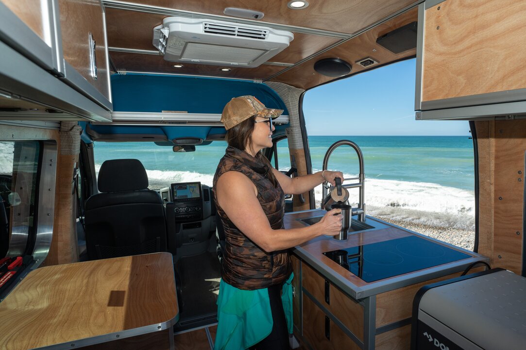 A woman enjoying a morning in her camper van kitchen, pouring coffee while admiring the ocean view. Experience serenity and convenience in our camper van.