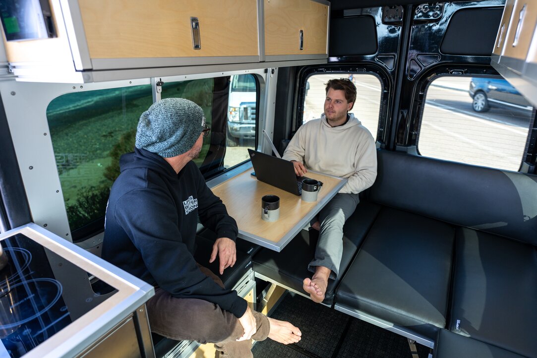 2 people inside of camper van having a conversation around a wall mounted fold down desk while sitting on sectional