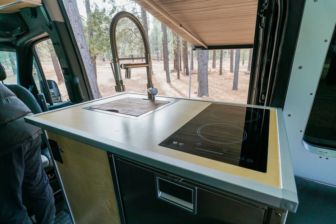 An all-in-one kitchen unit is pictured inside of the conversion van. The sink, faucet, stove, and refrigerator (Dometic CRX 80S) take the spotlight.