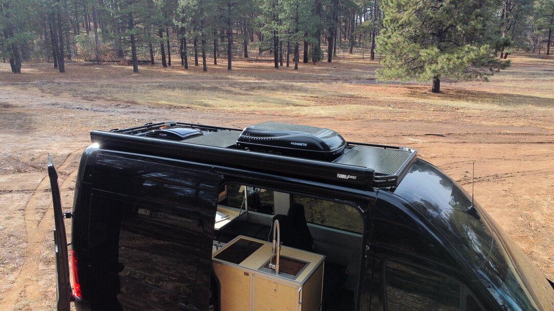 A roofrack sits atop the black Sprinter van showcasing many rooftop accessories. These include 200 watts of Ecoflow solar, a Dometic Penguin II heating and AC unit, a Maxx Fan and a Fiamma F45S awning