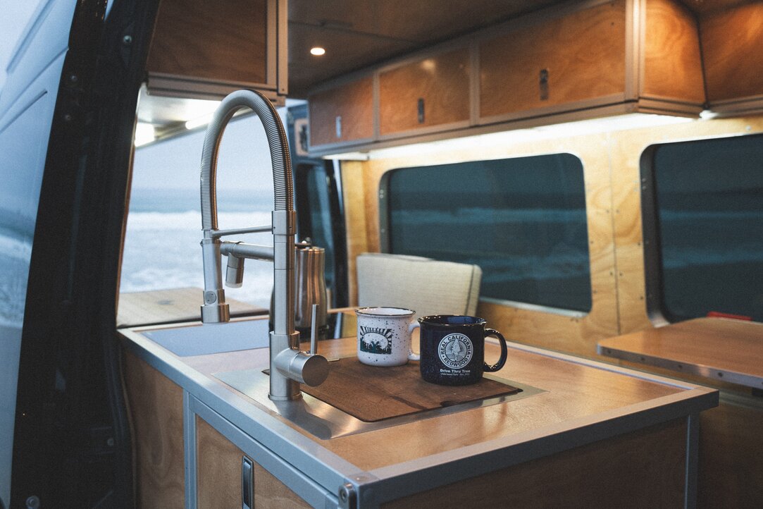 Explore the functional and stylish all-in-one kitchen counter. Coffee mugs, dual facets, and a cooktop with a coffee pot create a visually appealing setup against the backdrop of the camper van build.