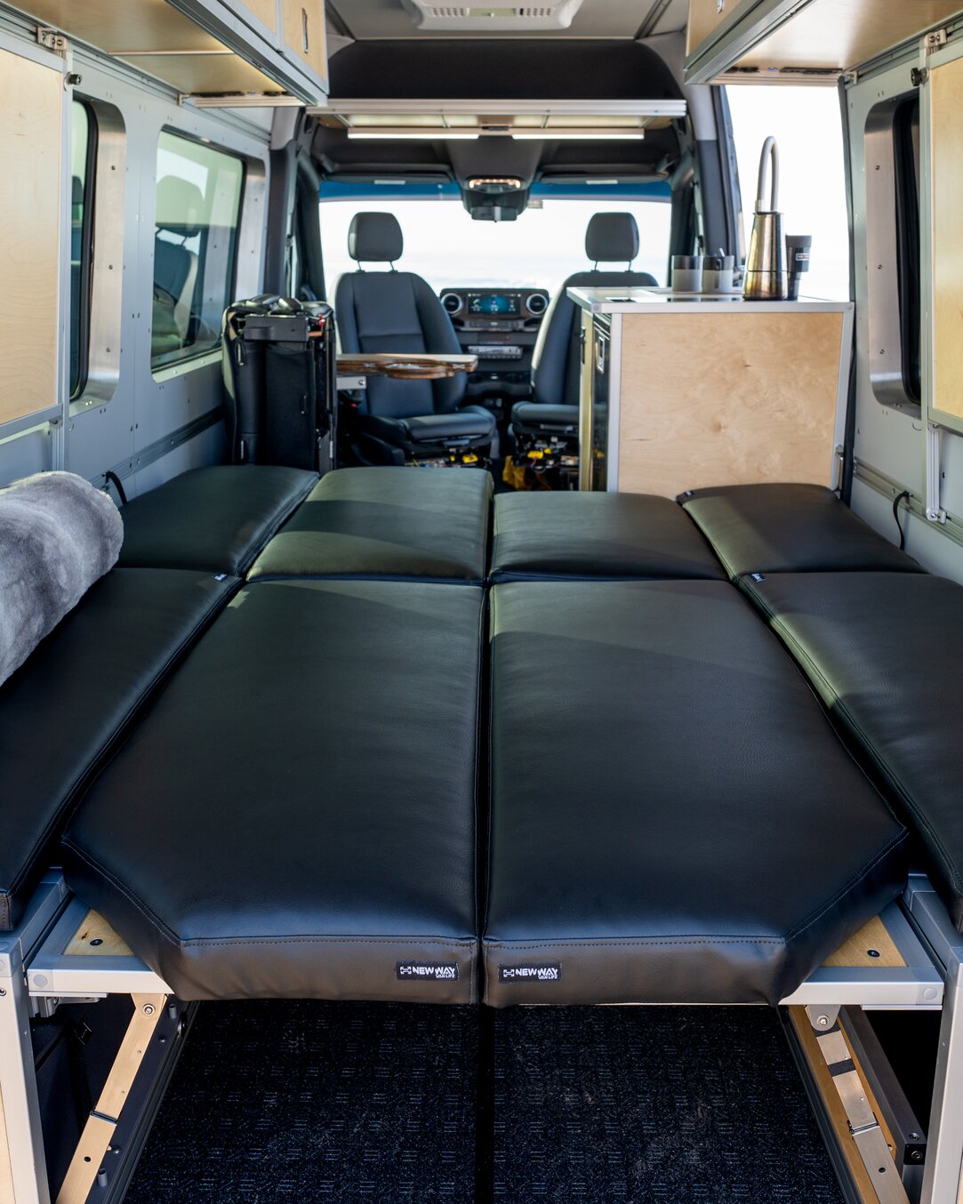 An almost queen-sized bed is made within this Sprinter 144 camper van. The bed is made with luxurious black cushions that are snuggly fit into place.