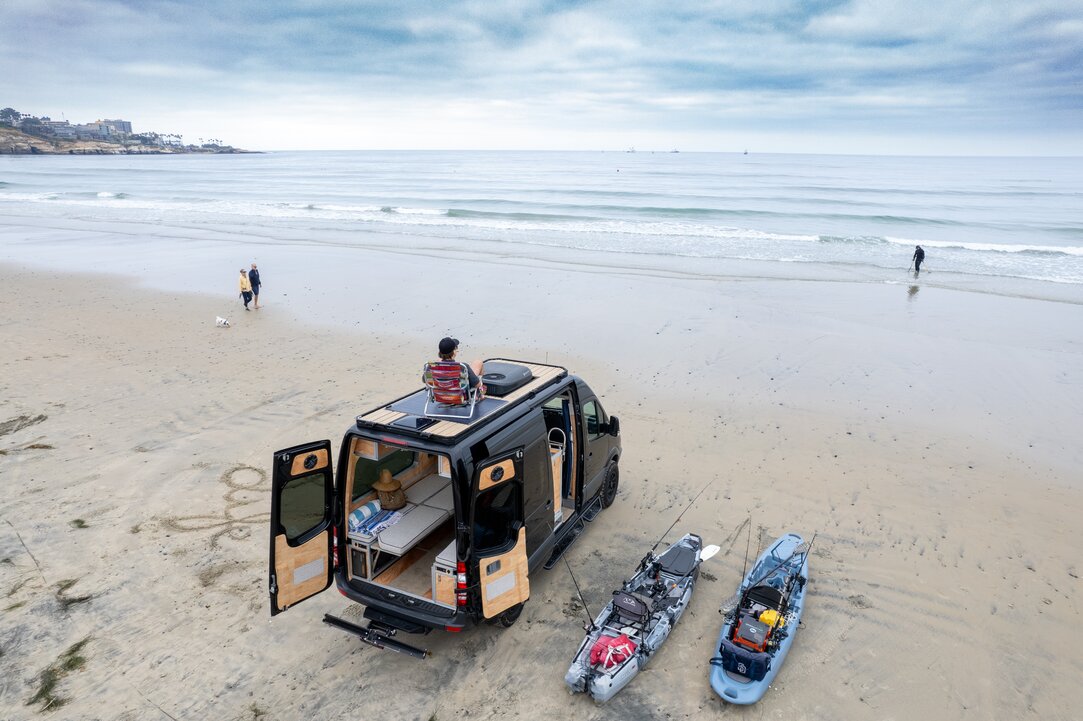 A woman enjoying a morning in her camper van kitchen, pouring coffee while admiring the ocean view. Experience serenity and convenience in our camper van.