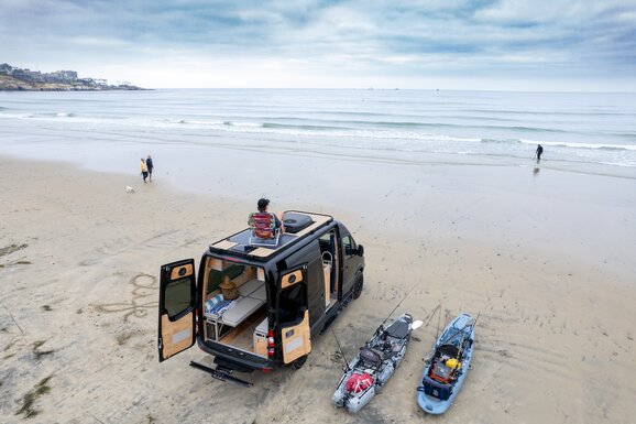 A woman enjoying a morning in her camper van kitchen, pouring coffee while admiring the ocean view. Experience serenity and convenience in our camper van.