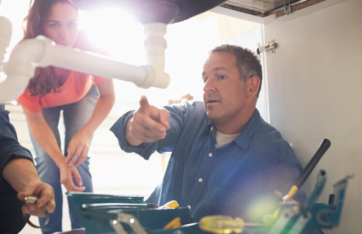 A contractor points to a pipe underneath a sink that needs to be fixed. A new homeowner is standing behind him.