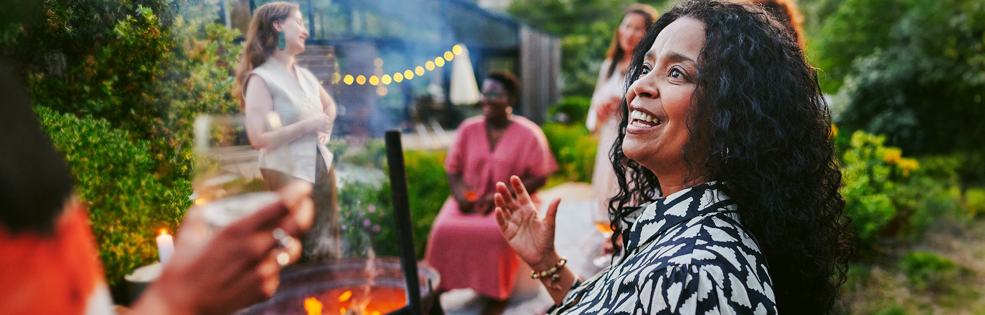 Woman with curly hair laughing joyfully at a backyard gathering with friends in a garden setting with string lights.