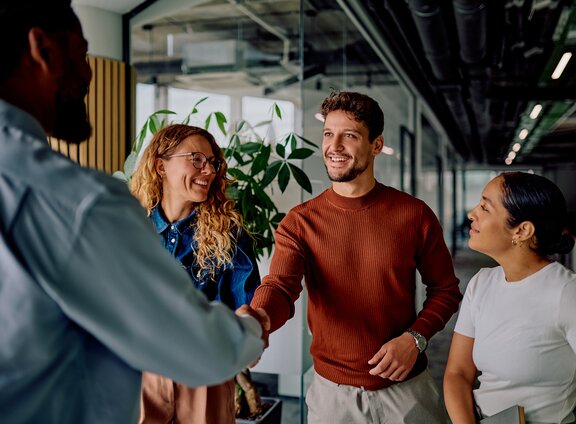 A group of diverse professionals smiling and shaking hands in a modern, sunlit office hallway.