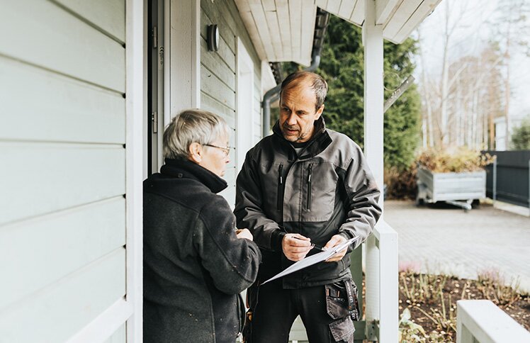 A man in a dark jacket holds papers and talks to an older woman in glasses on a porch.