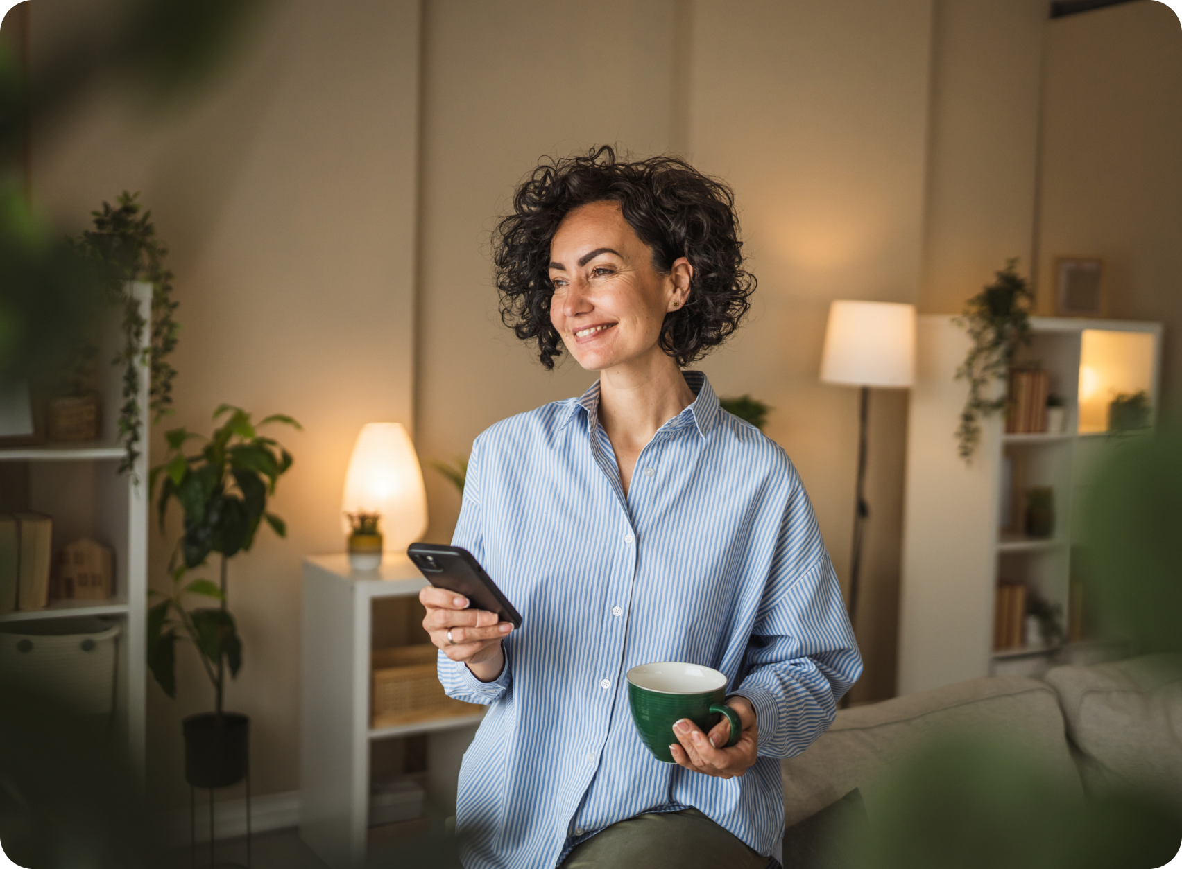 A woman with curly hair happily using her smartphone while holding a coffee mug at home. 