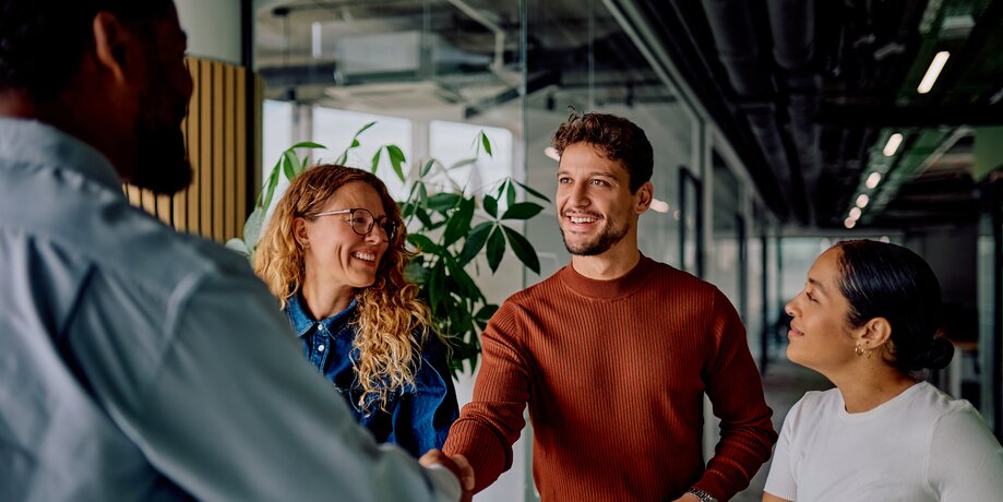 A group of diverse professionals smiling and shaking hands in a modern, sunlit office hallway.