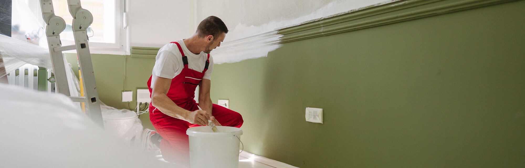 A man in red overalls painting a wall in a fixer-upper.