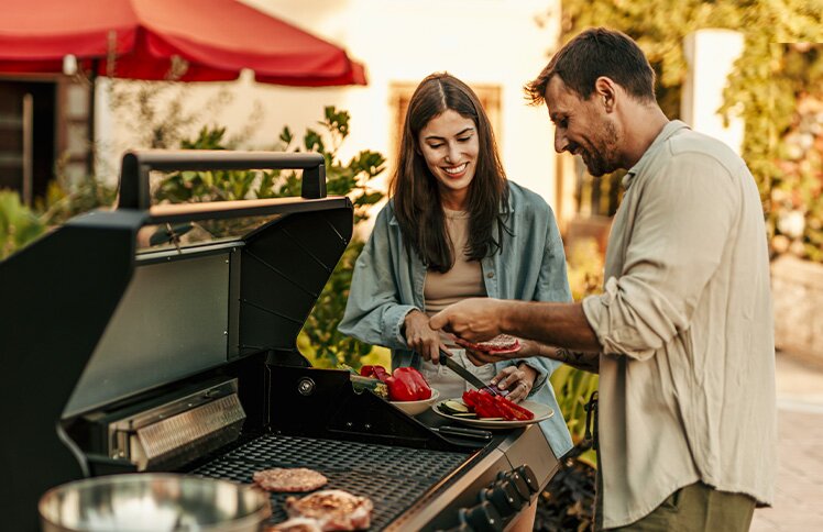 Two people stand smiling near an open grill, cooking a safe distance from their home.