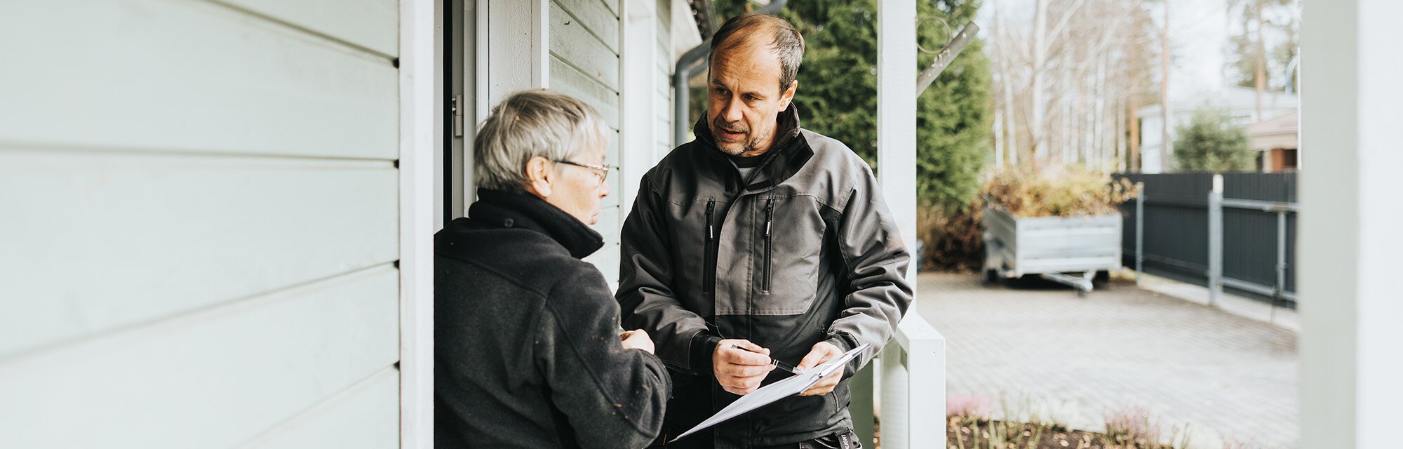 A man in a dark jacket holds papers and talks to an older woman in glasses on a porch.
