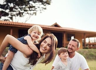 A family of four laughing together outdoors in front of a house, with a mother holding a young child on her back and a father sitting beside another child.
