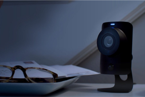 A small  home security camera standing on a table near glasses and documents