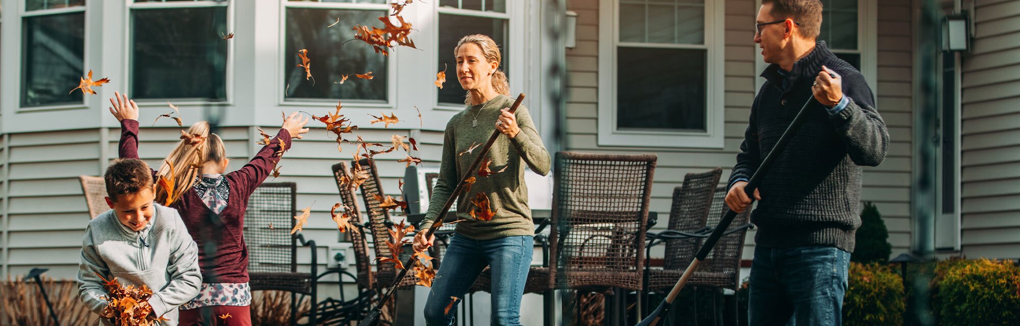 Two adult homeowners rake leaves in front of their home with two children.