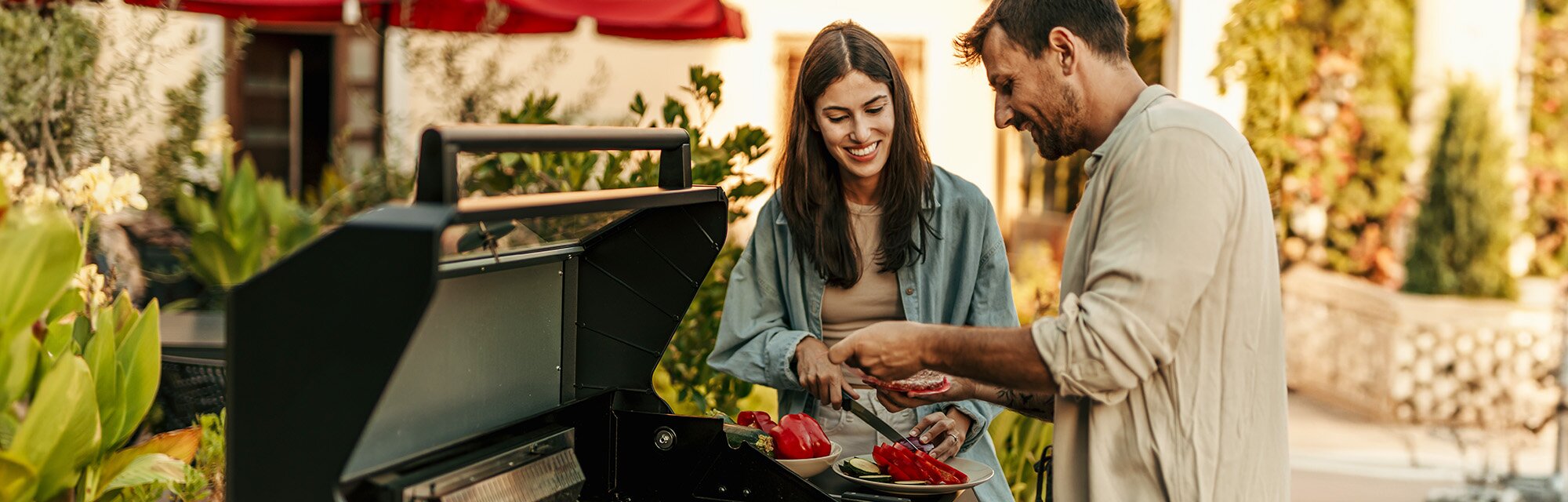 Two people stand smiling near an open grill, cooking a safe distance from their home.