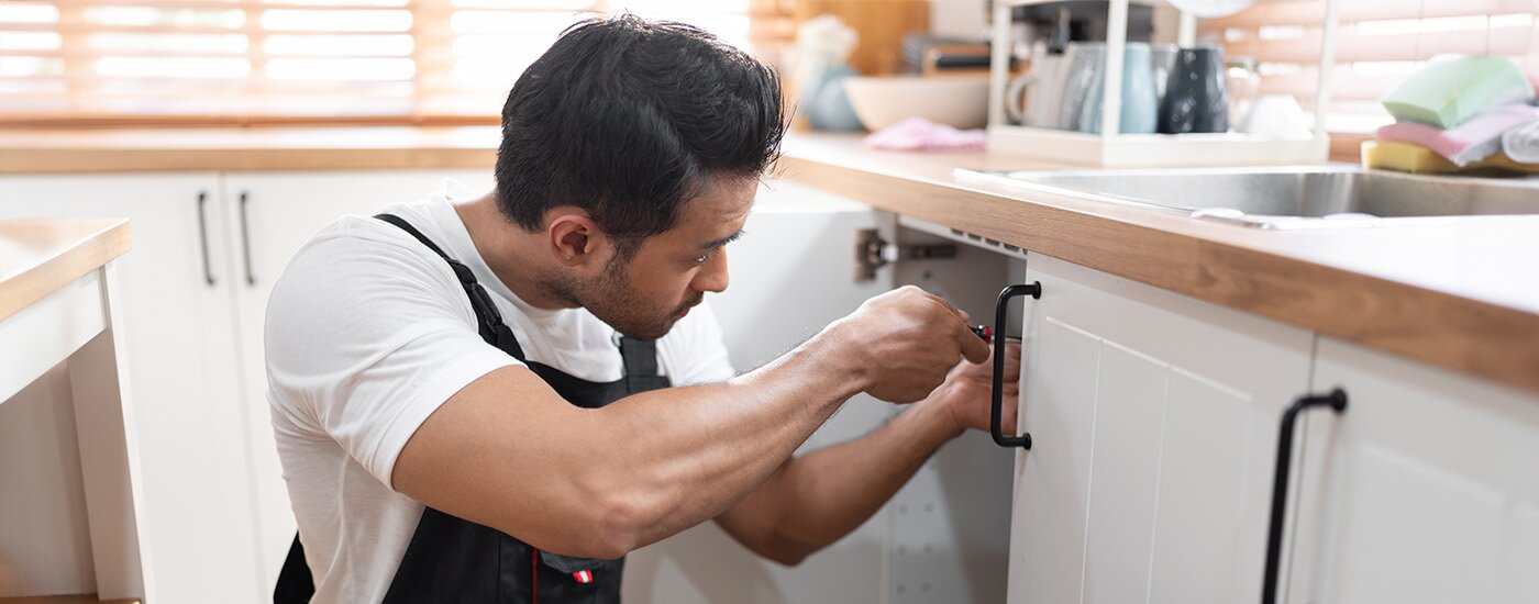 A homeowner wearing overalls performs maintenance on a kitchen sink while sun shines through the window blinds.