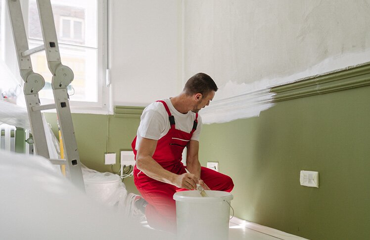 A man in red overalls painting a wall in a fixer-upper.